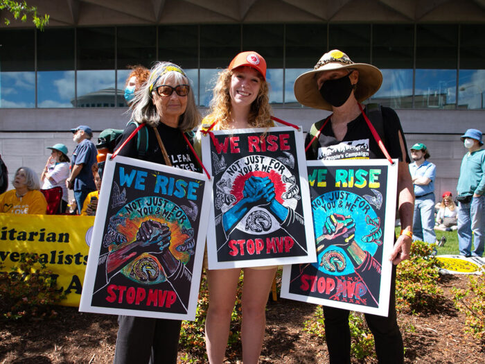 Three women wearing colorful protest signs pose at a rally to stop the Mountain Valley Pipeline.