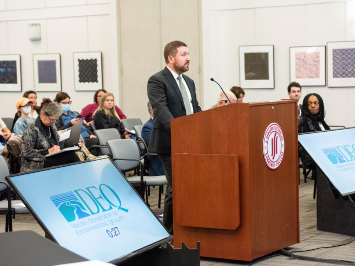 A serious-looking man in a suit stands at the podium in a crowded public meeting room.